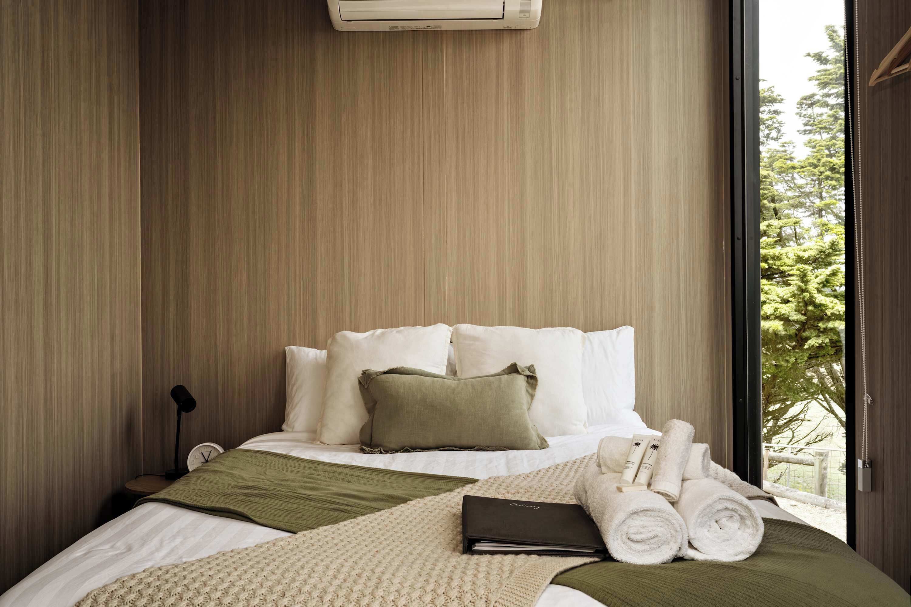 Una cama bien hecha con ropa de cama blanca y verde, una lámpara de noche y una ventana con vistas a árboles.