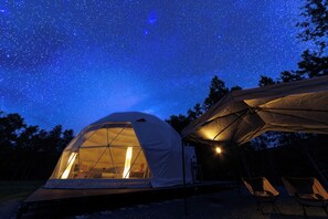 Una tienda de forma abovedada con vistas al cielo estrellado, otra tienda más pequeña y dos sillas afuera.