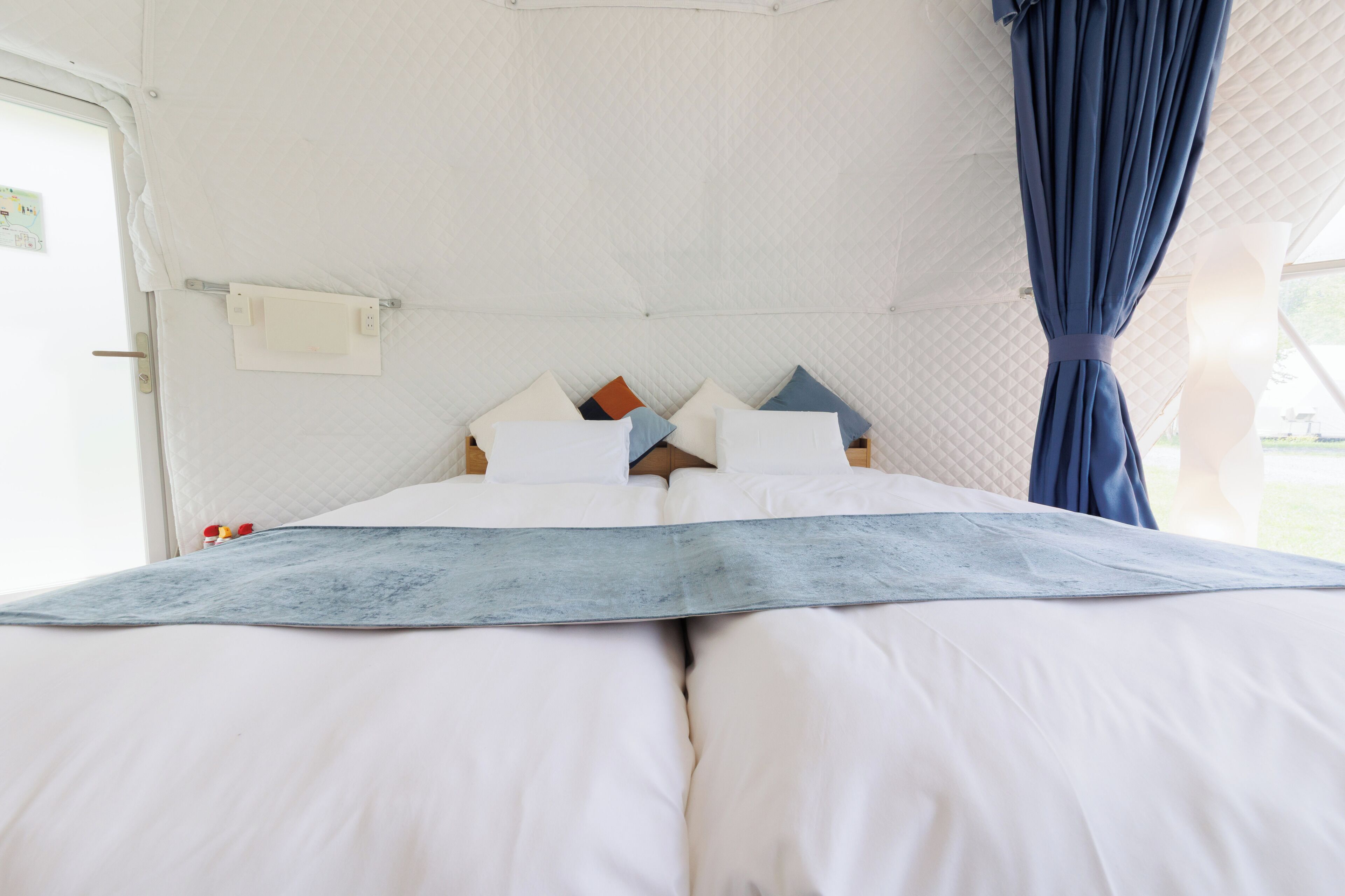 Una cama con ropa de cama blanca y una manta azul, cabecera con varios almohadones y una cortina al lado derecho.