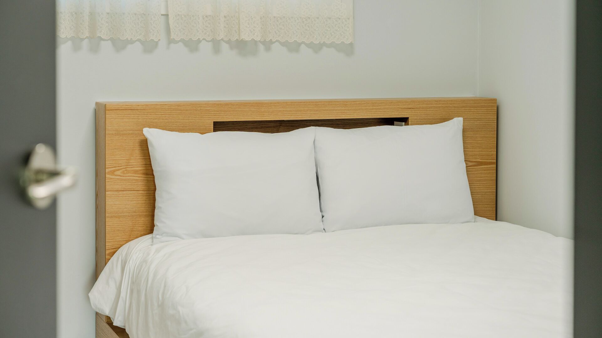 A bed with a wooden headboard and white bedding in a room with a door and a window with a lace curtain.