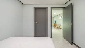 Une chambre à coucher avec un lit, une porte fermée et une porte ouverte donnant sur un couloir où se trouvent un banc et un téléviseur.