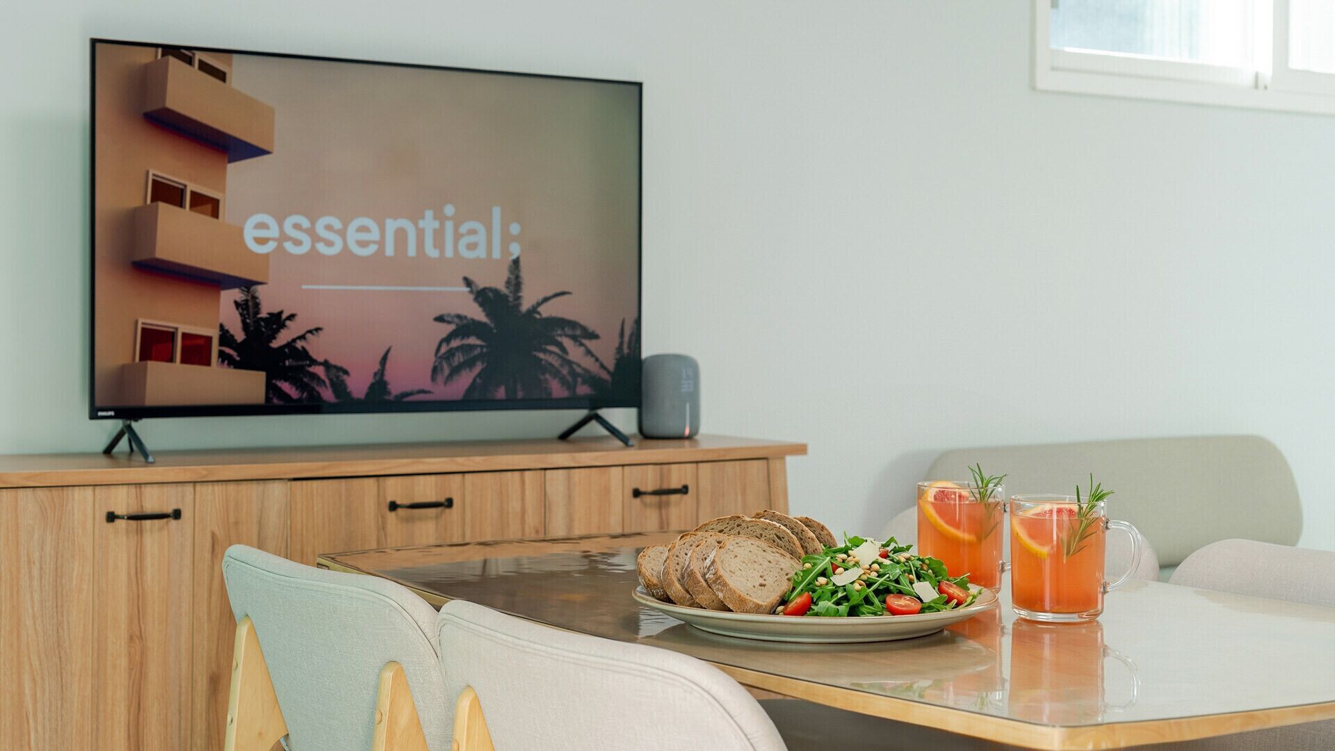 A modern living room with a flat-screen TV, wooden furniture, a glass table, and a plate of food and drinks.