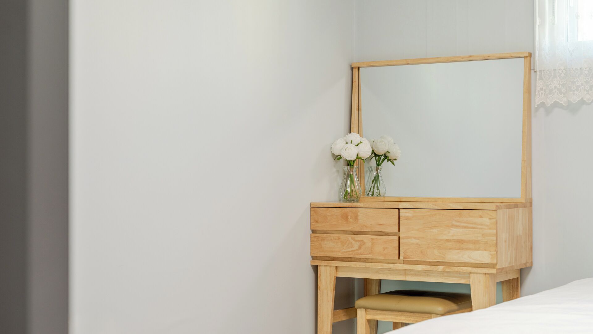 A wooden dresser with a mirror, a vase with white flowers, and a bench in a room with a plain wall and a window with white curtains.