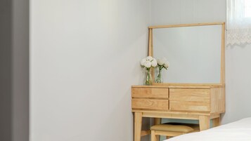 Un aparador de madera con espejo, un jarrón con flores blancas y un banco en una habitación con pared lisa y ventana con cortinas blancas.