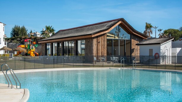 A swimming pool with a wooden building in the background, a fenced area, and a clear sky. - Exclusive Penthouse in Paraíso Pueblo – Style & Serenity (Benahavís)