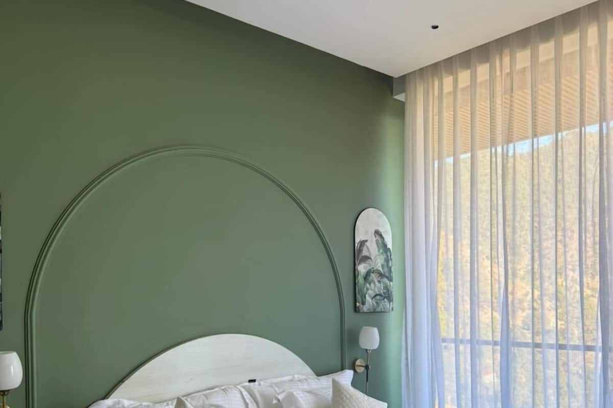 A neatly made bed with white bedding and pillows, a green wall, and a window with sheer curtains.