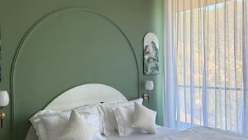 A neatly made bed with white bedding and pillows, a green wall, and a window with sheer curtains.