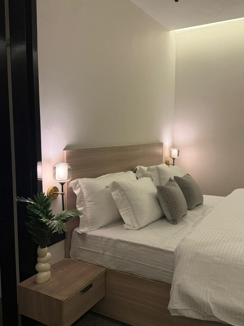 Ein ordentlich bezogenes Bett mit vielen Kissen, ein Nachttisch mit Lampe und ein kleiner Blumentopf.