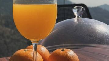 Um copo de suco de laranja, uma laranja inteira e meia laranja sobre uma mesa, com vista para montanhas ao fundo.
