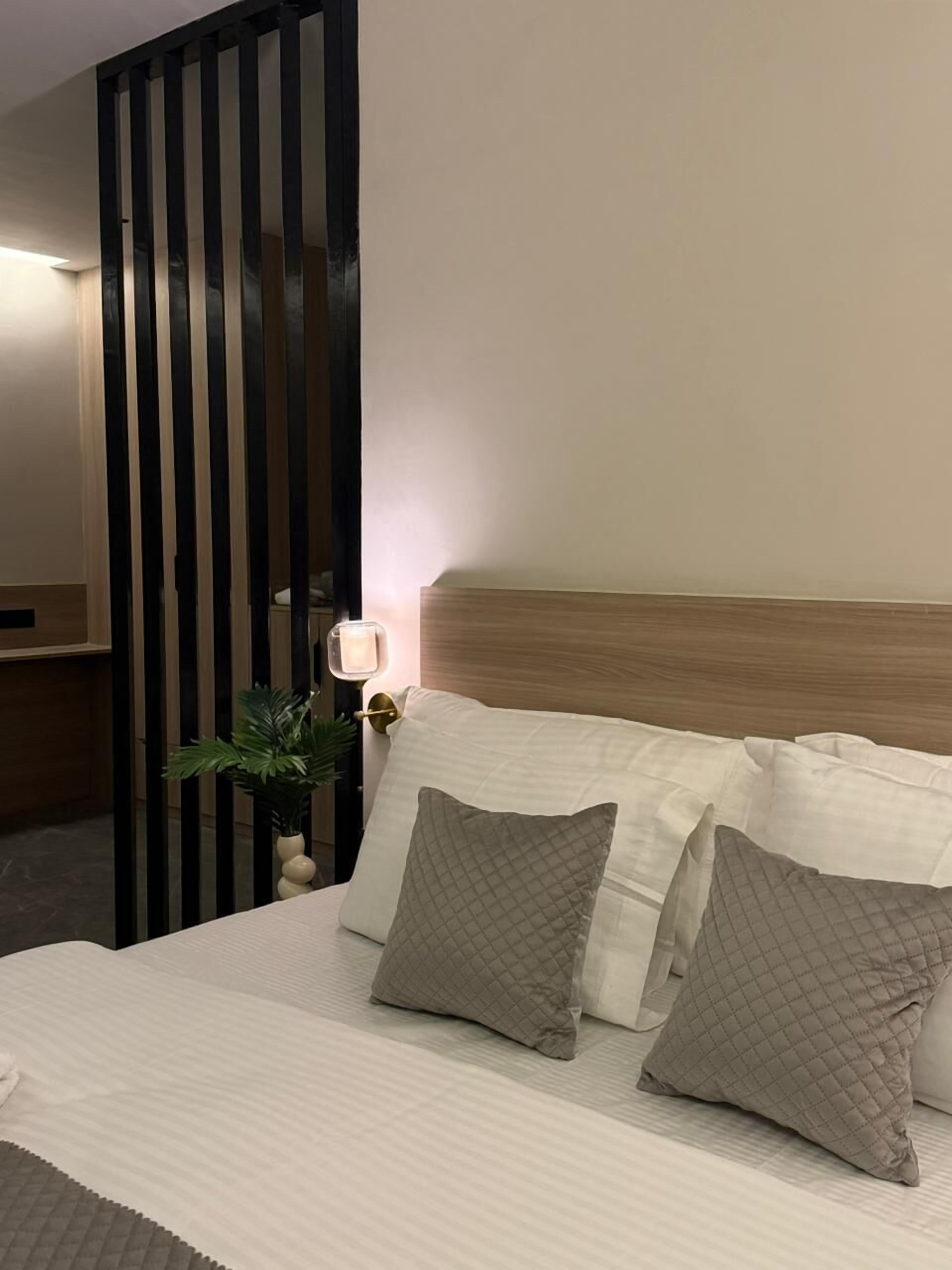 A hotel room with a bed, two pillows, a nightstand with a lamp, and a plant.