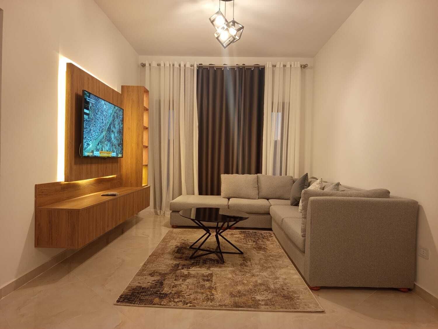 Ein modernes Wohnzimmer mit einem an einer Holzplatte montierten Flachbildfernseher, einem Ecksofa, einem Couchtisch und einem gemusterten Teppich.