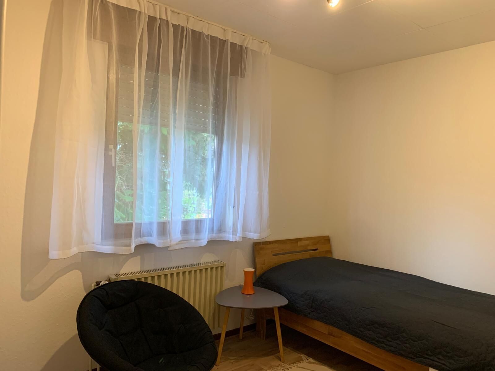 A small, single-bed room with a wooden headboard, a round table with a cup, a black chair, and sheer curtains over a window.