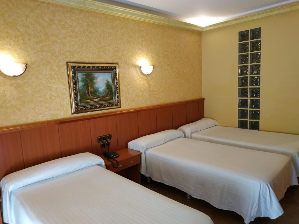 A hotel room with three single beds, a wooden headboard, a painting on the wall, and a window with blinds.