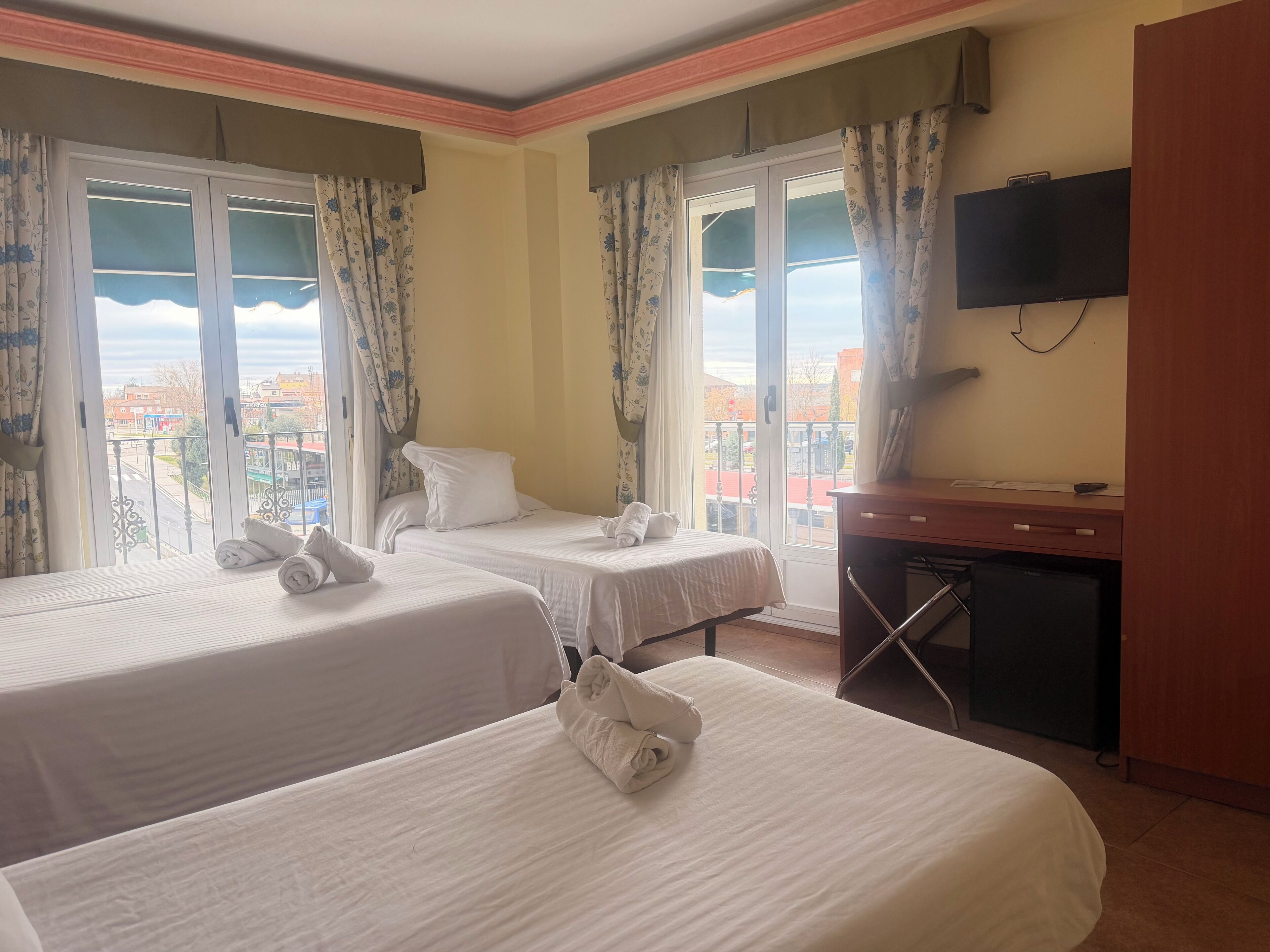A hotel room with two beds, a desk, a TV, and a balcony with a city view.