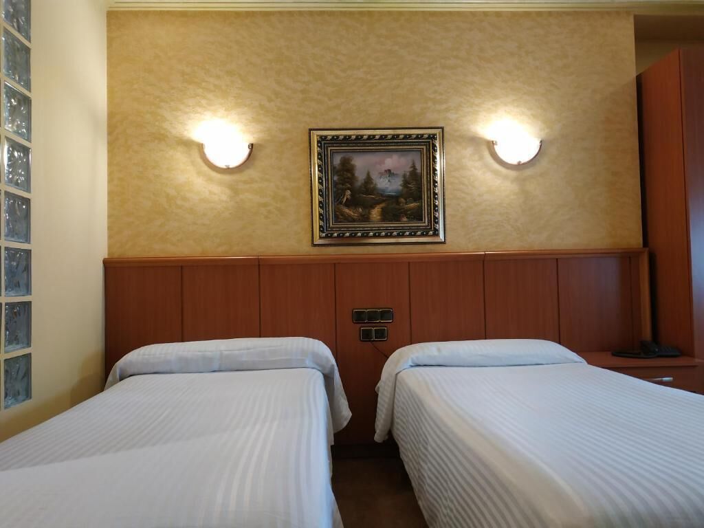 A hotel room with two single beds, a framed picture on the wall, and two wall-mounted light fixtures.
