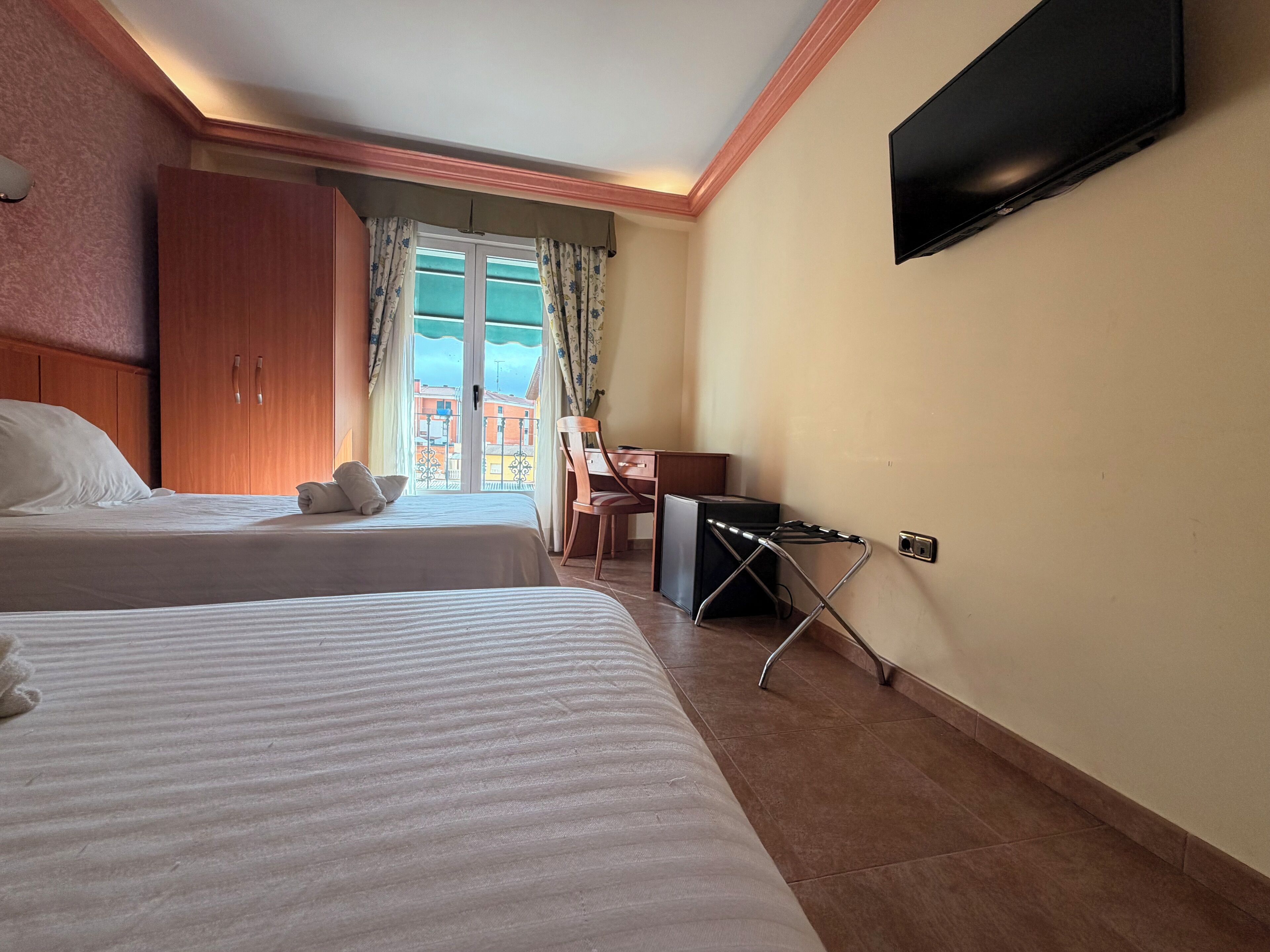 Habitación de hotel con dos camas, un escritorio, una silla, un televisor y vistas a edificios a través de una ventana.