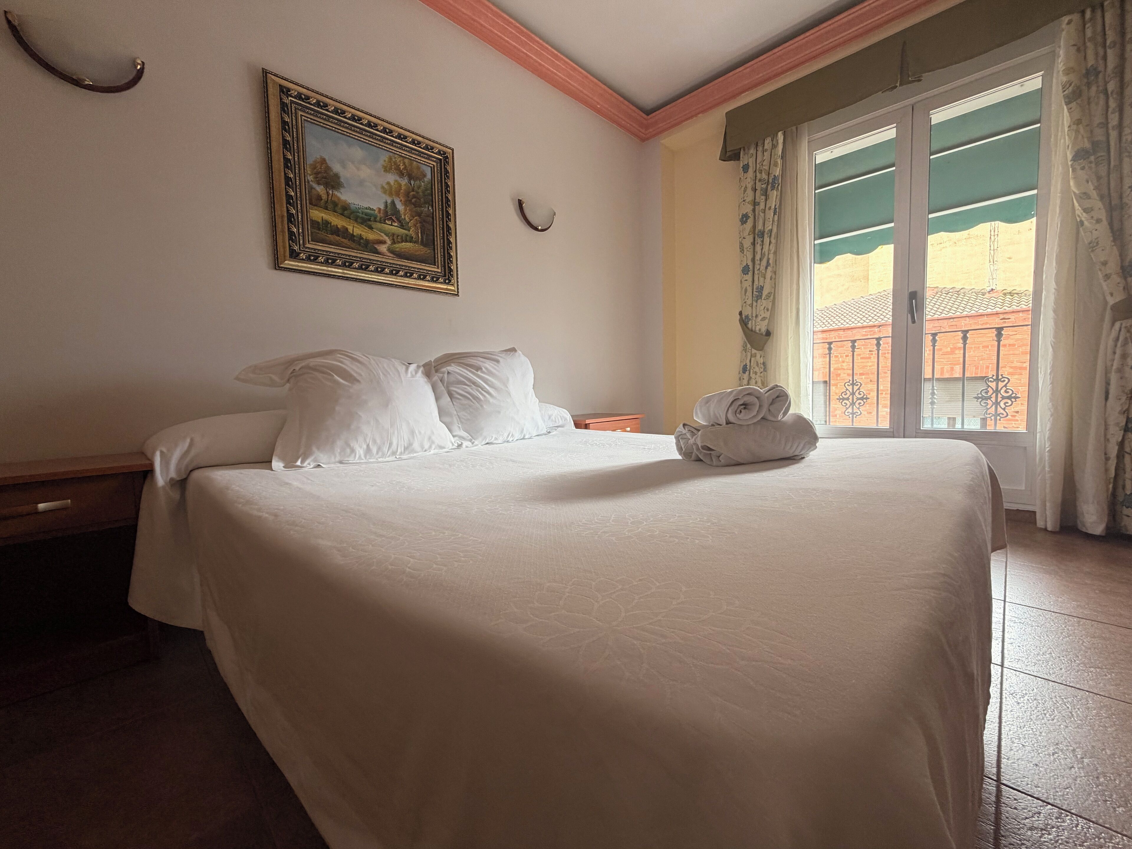 A neatly made bed with white linens and pillows, a painting on the wall, and a window with curtains.