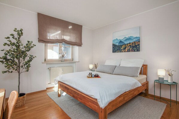 A bedroom with a wooden bed, a window with a brown valance, a bedside table with a lamp, a wall-mounted painting, and a potted plant.