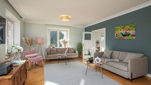 Ett rymligt vardagsrum med en grå soffa, en beige soffa och ett soffbord i trä. Ett stort fönster släpper in naturligt ljus, och på väggen hänger en färgstark, abstrakt tavla.