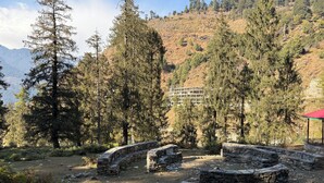 A mountainous area with stone ruins, trees, and a building in the background. - Manali Se - Your Home in the Pines (Jagatsukh)