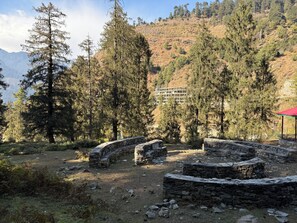 A mountainous area with stone ruins, trees, and a building in the background. - Manali Se - Your Home in the Pines (Jagatsukh)