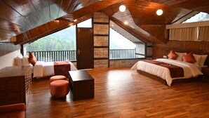 A spacious room with a wooden ceiling, a large bed, a seating area with a table, and a view of greenery outside. - Manali Se - Your Home in the Pines (Jagatsukh)