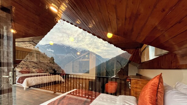 A bedroom with a bed, pillows, a wooden ceiling, and a mountain view through a window. - Manali Se - Your Home in the Pines (Jagatsukh)