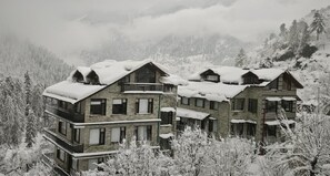A multi-story building with balconies, surrounded by snow-covered trees and mountains. - Manali Se - Your Home in the Pines (Jagatsukh)