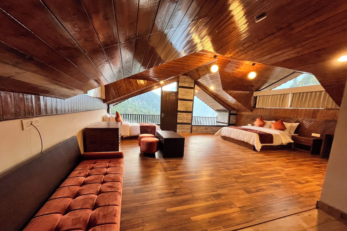 A spacious attic bedroom with a wooden ceiling, a large window, a sofa, a bed, and a small table.