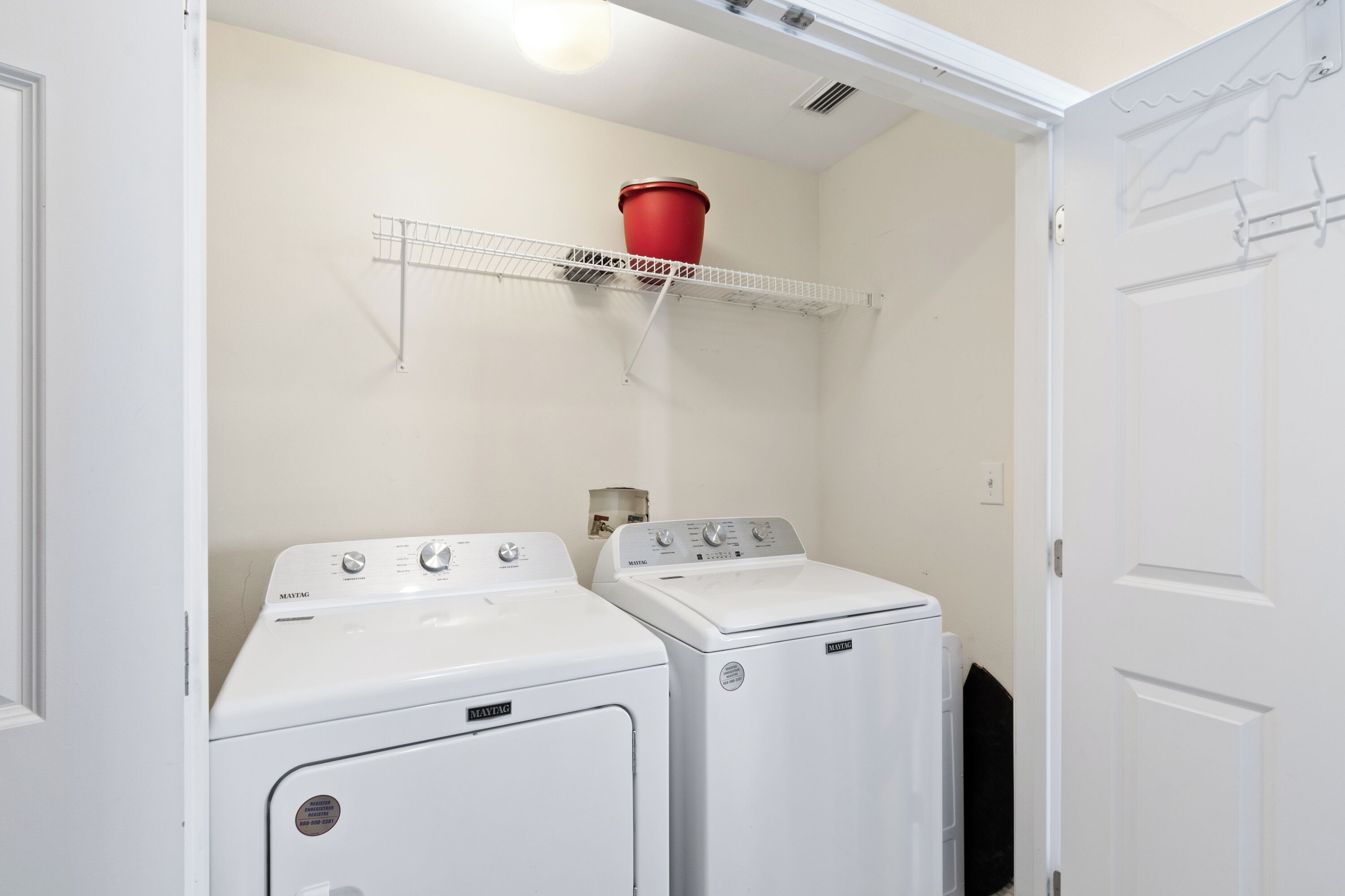 A laundry room with two white front-loading washers and dryers, a metal drying rack, and a red bucket on the wall.