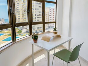 Una habitación con vista a una piscina y edificios, una mesa blanca con una planta y una silla verde.