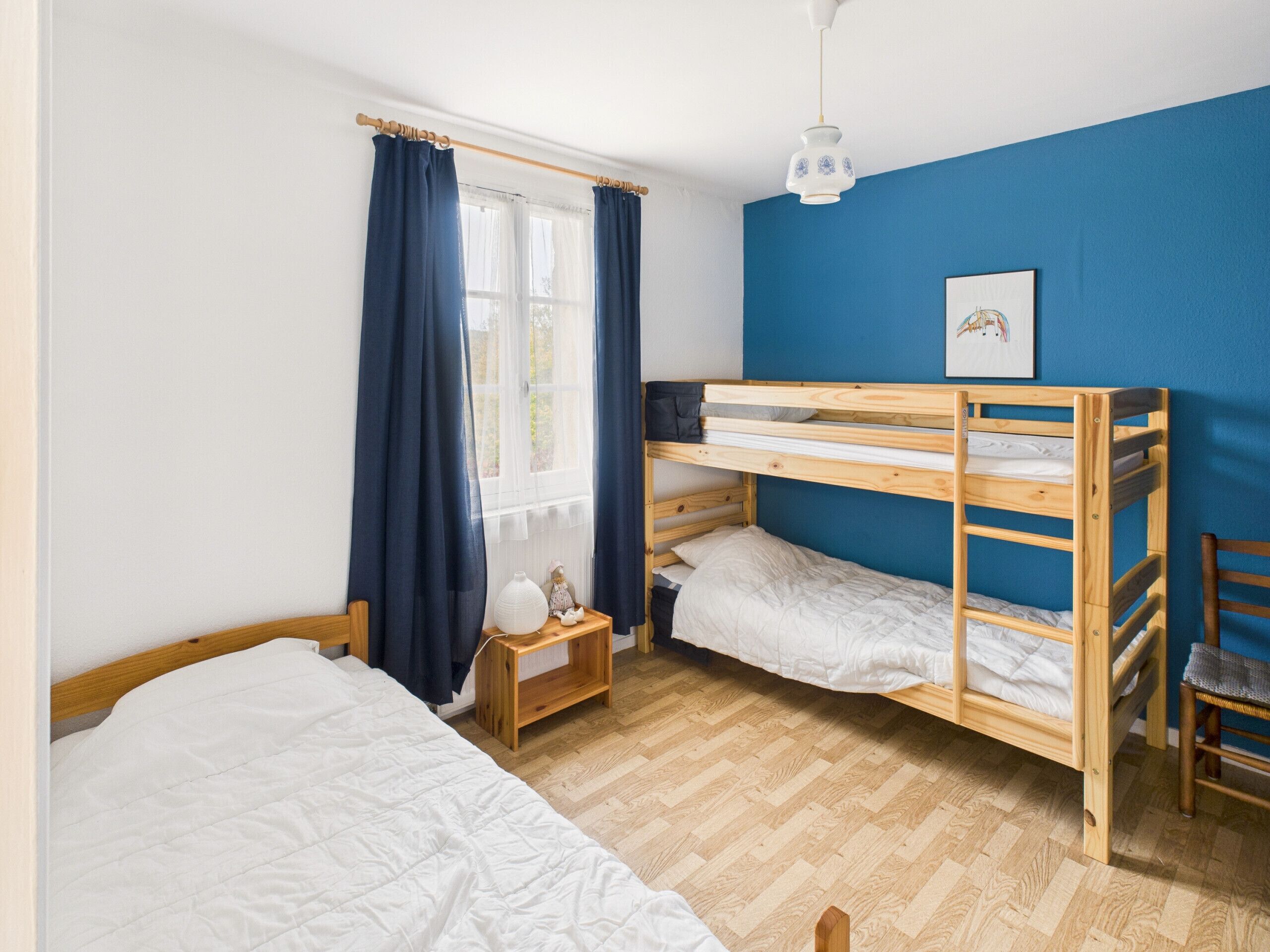 Ein Zimmer mit zwei Etagenbetten, einem kleinen Nachttisch mit Lampe, einem Fenster mit Vorhängen und einer blauen Wand.