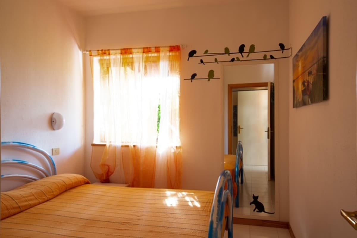Une chambre à coucher avec un lit, une fenêtre avec des rideaux, une porte, un miroir et une décoration représentant un oiseau fixée au mur.