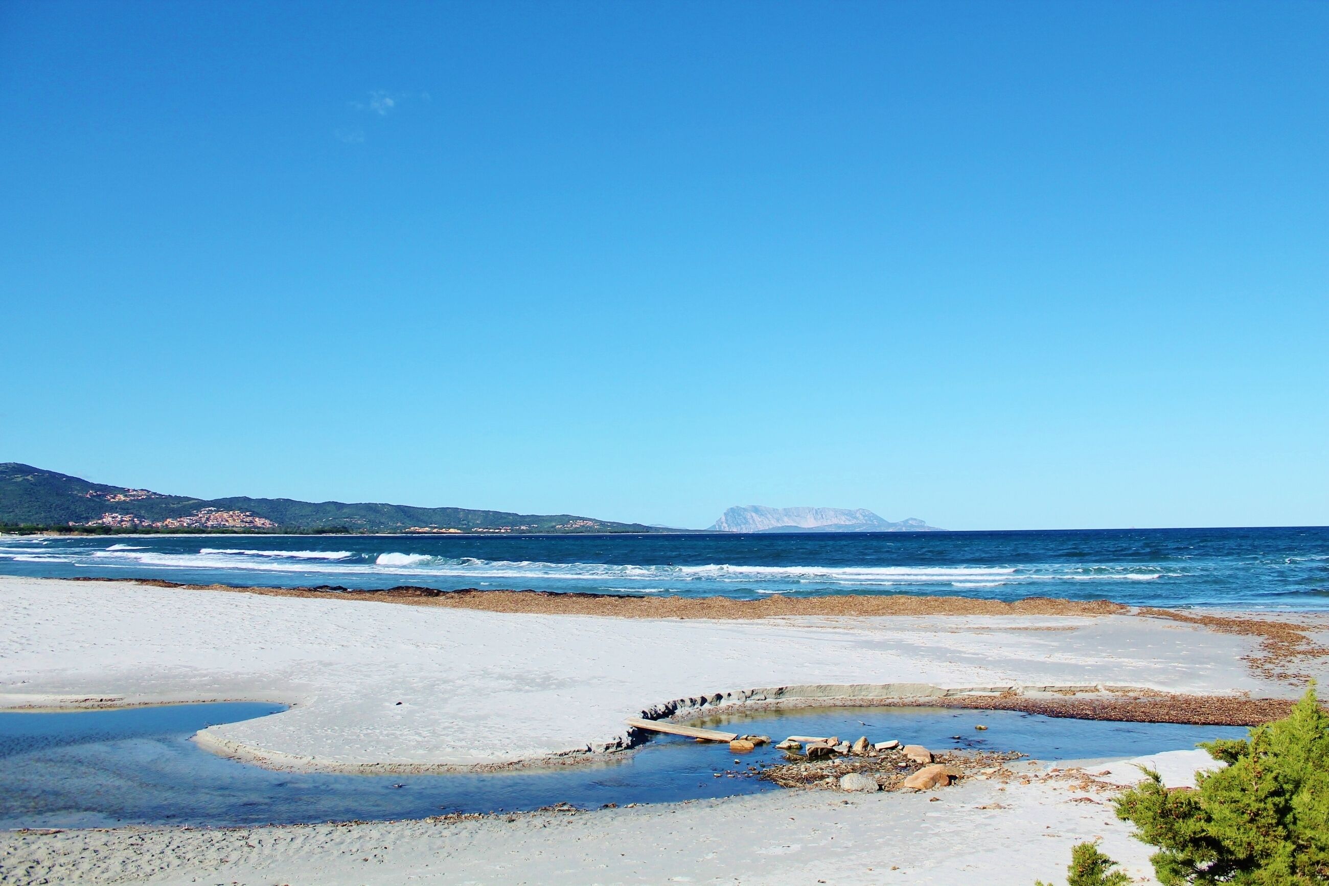 A beach with a rocky shoreline, clear blue sky, and distant mountains.