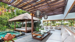 Eine Holzterrasse mit einem Glastisch, einem Gazebo mit Strohdach, einem Pool und einem weißen Sofa mit Kissen.