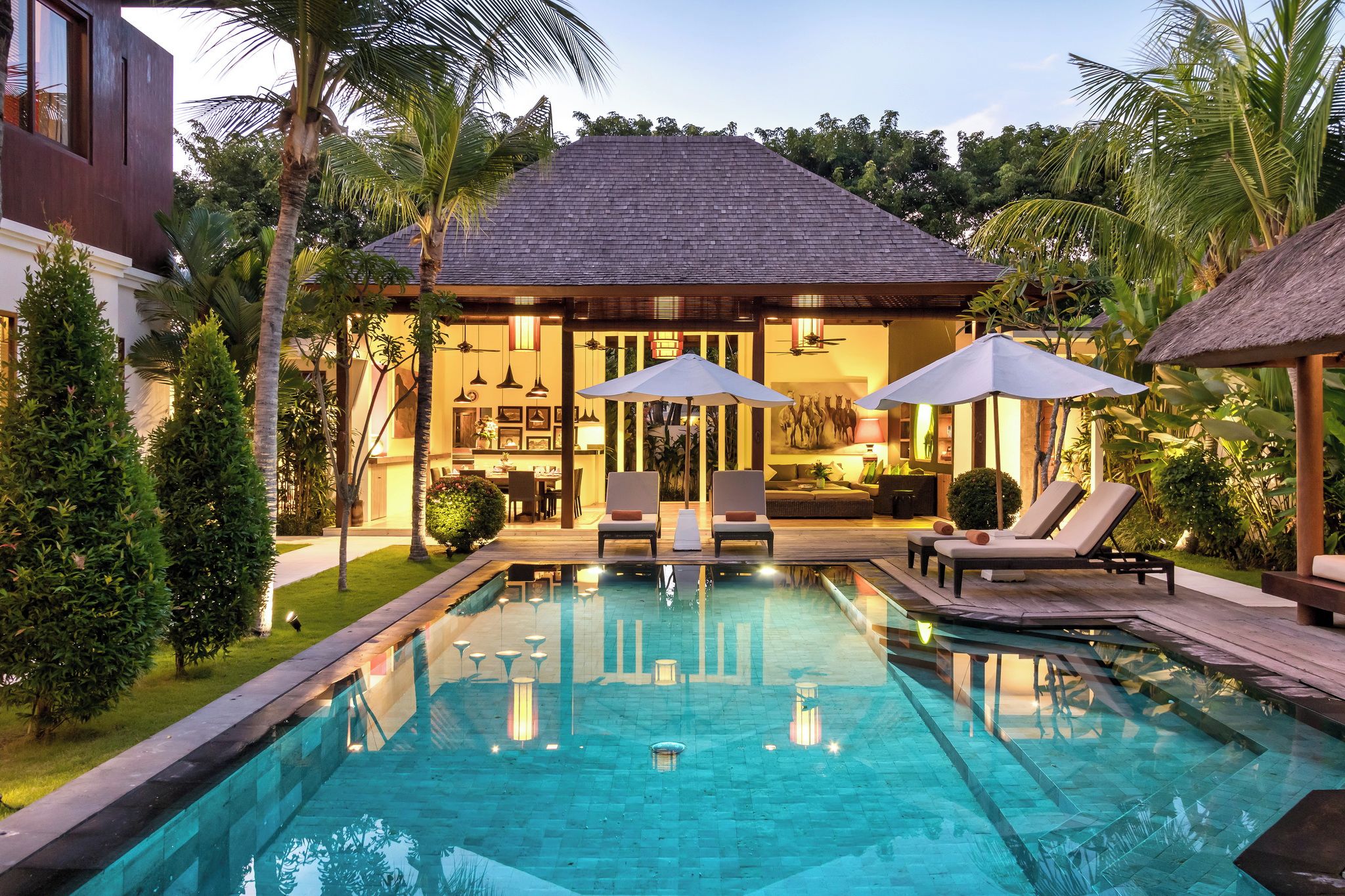 A pool area with lounge chairs and umbrellas, a dining area with a kitchen, and a building with a tiled roof.