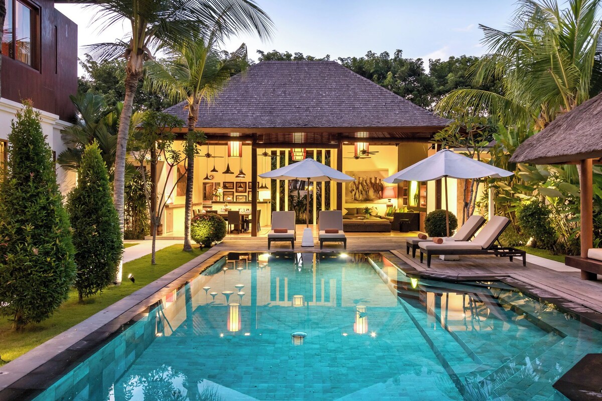A pool area with lounge chairs and umbrellas, a dining area with a kitchen, and a building with a tiled roof.