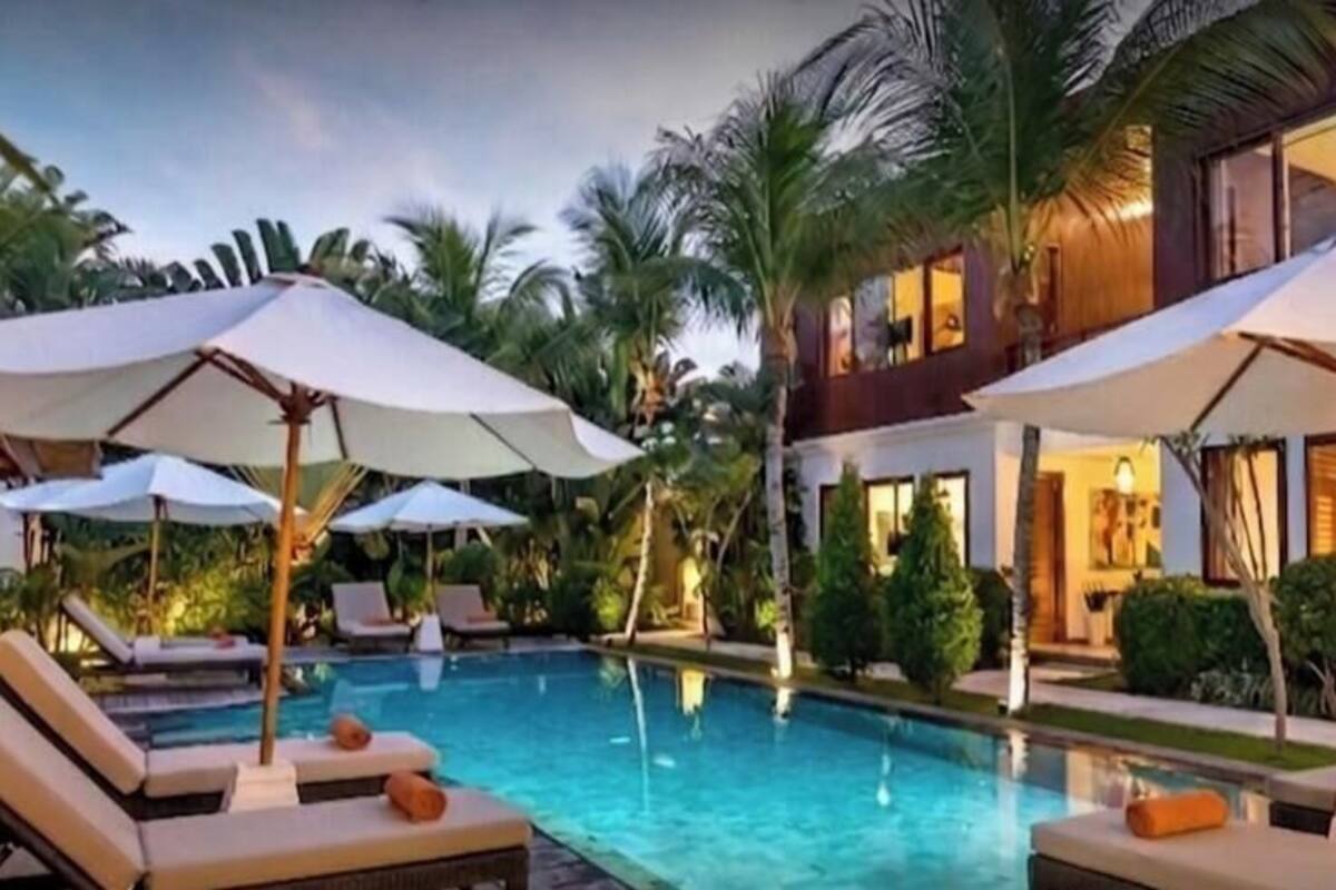 A pool area with lounge chairs and umbrellas, surrounded by palm trees and a building with multiple windows.