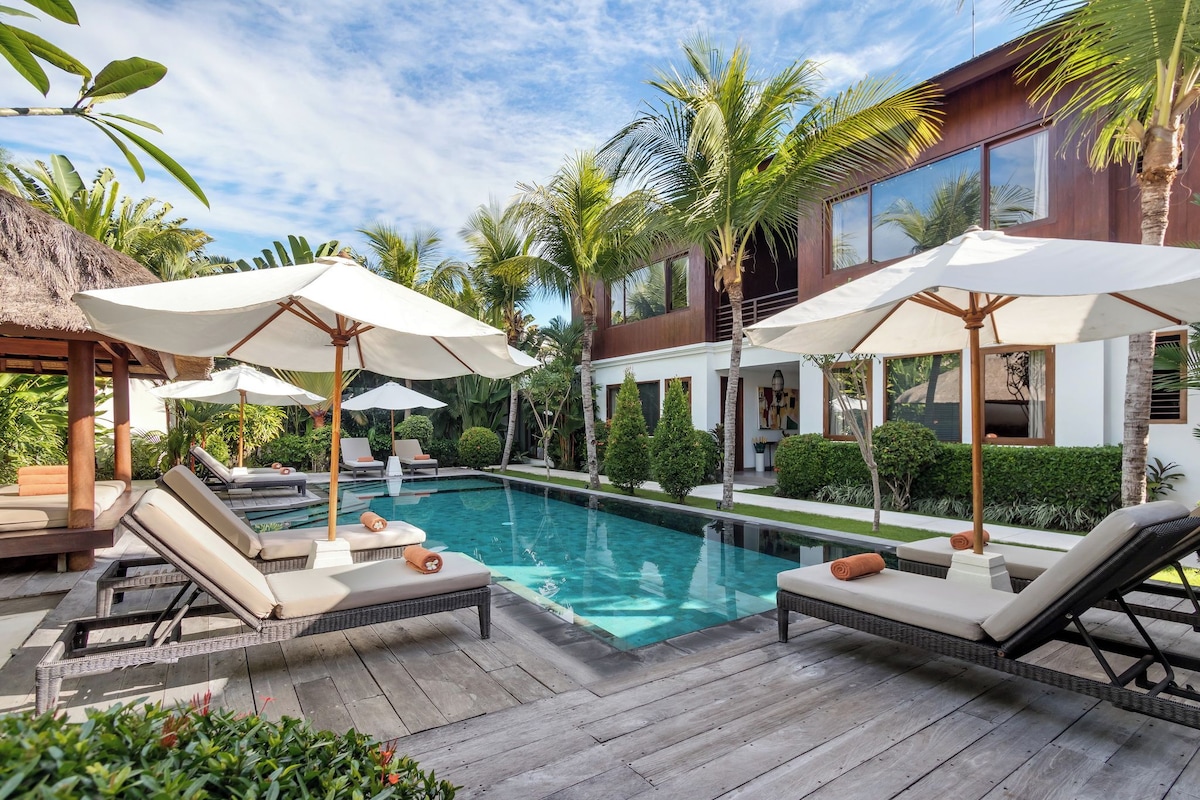 A poolside area with lounge chairs and umbrellas, surrounded by palm trees and a modern building.