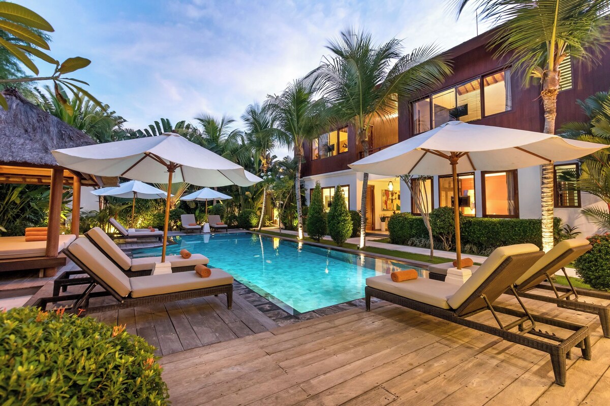 A poolside area with lounge chairs and umbrellas, surrounded by palm trees and a modern building.