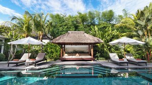 Un espace aménagé au bord de la piscine, avec des chaises longues, un gazebo à toit de chaume et une végétation tropicale luxuriante.