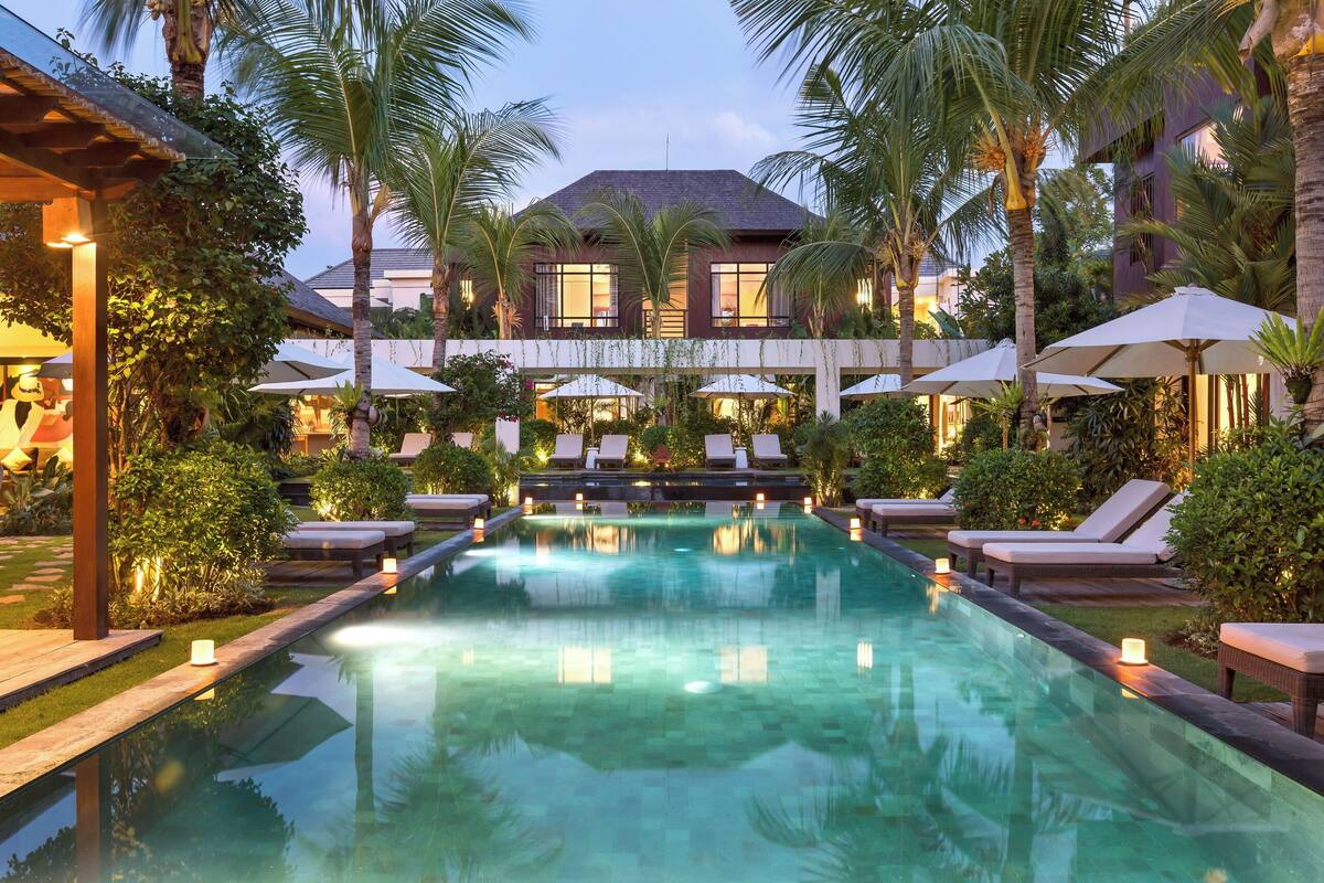 A pool area with lounge chairs and umbrellas, surrounded by palm trees and a building with large windows.