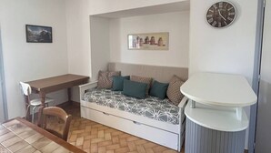 A compact living space with a dining table, a sofa with patterned cushions, a small white table, and a wall clock. - Les Mésanges Bleues (Le Poët-Laval)