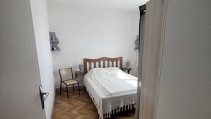 A bedroom with a wooden herringbone floor, a bed with a white bedspread, a chair, bedside tables with lamps, and a ceiling fan. - Les Mésanges Bleues (Le Poët-Laval)
