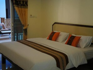 A bed with white bedding and orange pillows, a wooden headboard, and a view of a balcony with a person sitting outside. - Manee Guest House (Chiang Mai)