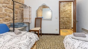 Une chambre avec un mur en pierre, une chaise en bois, un lit recouvert d’une couvre-lit à motifs, un porte-serviettes suspendu et un miroir.