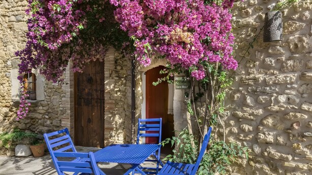 Un bâtiment en pierre avec une porte en bois, une table et des chaises bleues, et des fleurs de bougainvillier roses.