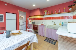A kitchen with a dining table, chairs, a refrigerator, a sink, and a shelf with kitchen items.