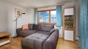 A modern living room with a large sectional sofa, a floor lamp, a wooden coffee table, and a view of a mountain through a window with blue curtains. - 1177 House Rosa & Alfred (Gaschurn)