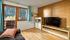 A modern room with a large window, wooden floor, a sofa with a cushion, a TV on a wooden stand, and a small shelf with decorative items. - 1177 House Rosa & Alfred (Gaschurn)